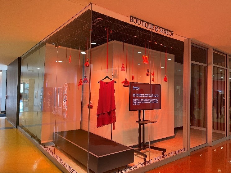 A display case with a hanging red dress surrounded by hanging paper dresses. A screen with event details.