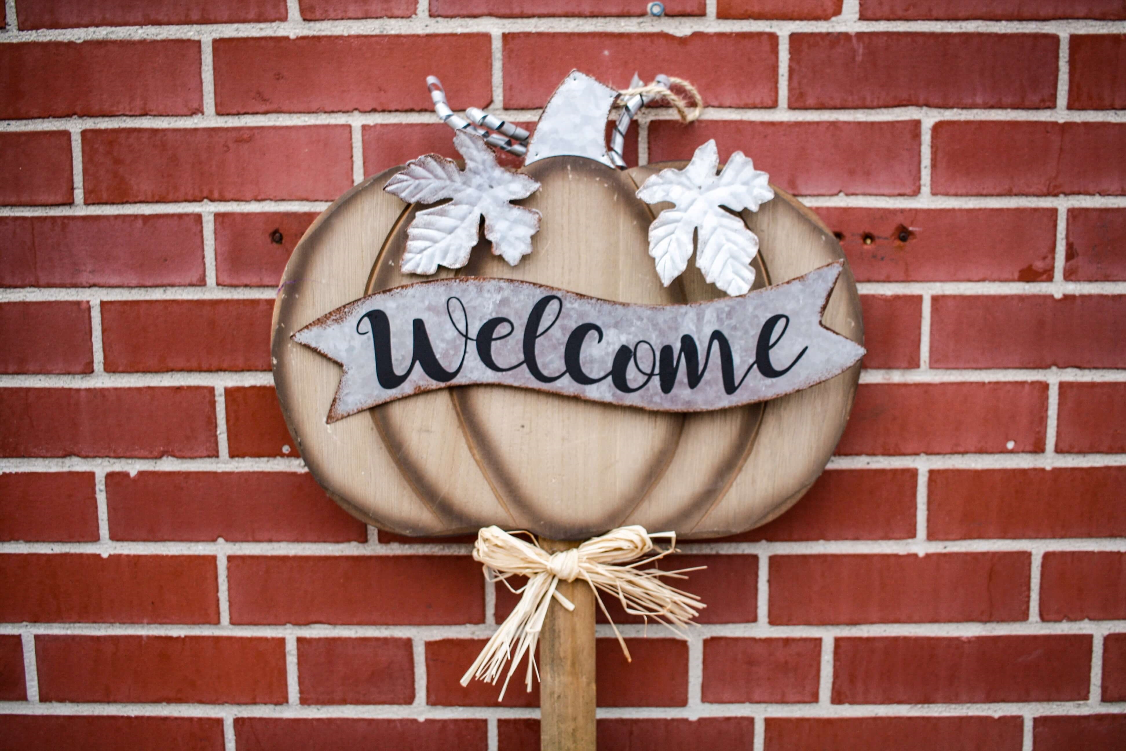 A brown and gray pumpkin-shaped signed with a "Welcome" banner on it hanging on a red brick wall